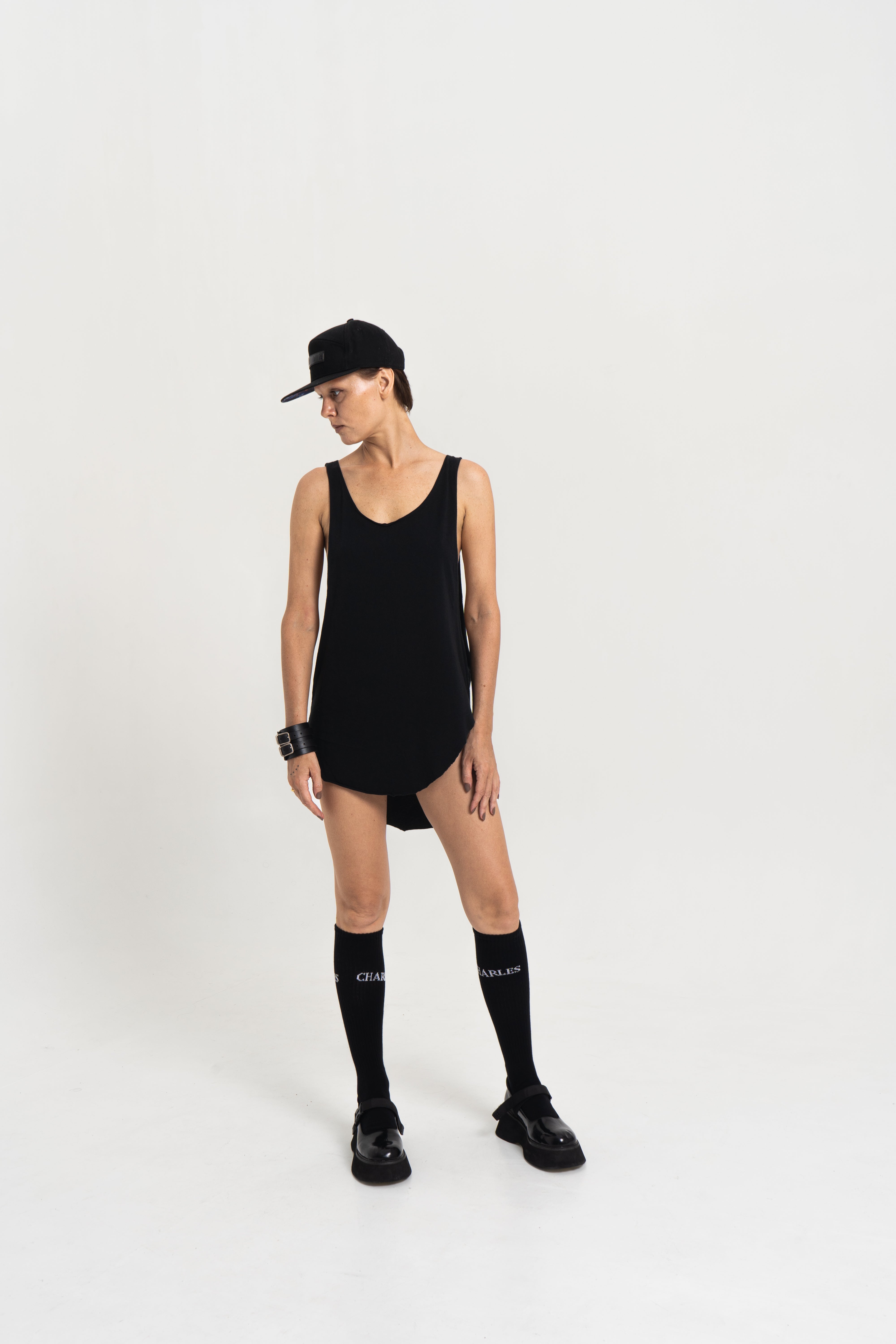 Model wearing the Charles Darius deep crew-neck tank in black, styled with high socks and black shoes, standing against a white studio background.